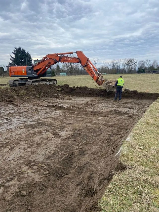 Koparka gąsienicowa zdejmuje warstwę humusu pod płytę fundamentową, pracownik wyznacza krawędź wykopu