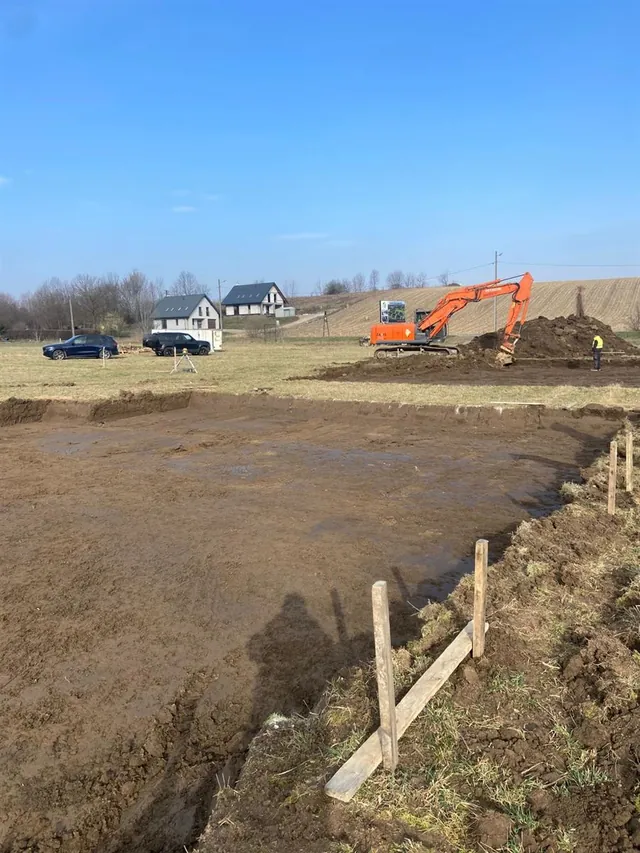 Wyrównany wykop pod pierwszy budynek z wbitymi palikami ciesielskimi, w tle koparka i istniejąca zabudowa sąsiedztwa