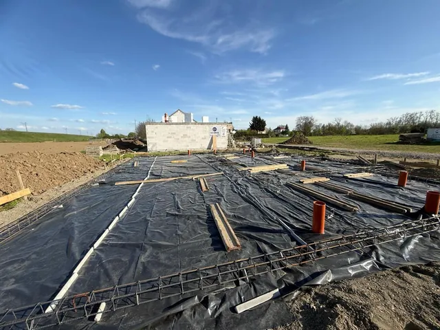 Zbrojenie płyty fundamentowej kolejnego budynku ułożone na folii izolacyjnej, z wyprowadzonymi pionami kanalizacji, w tle gotowe ściany sąsiedniego budynku dwulokalowego