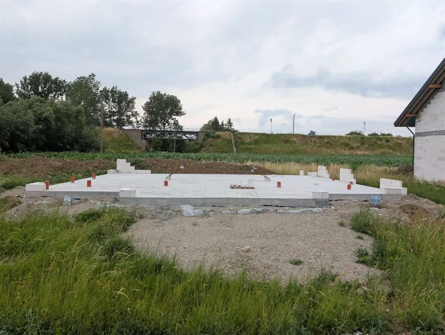 Płyta fundamentowa następnego budynku dwulokalowego z wypuszczonym zbrojeniem startowym, obok ukończony budynek z dachem