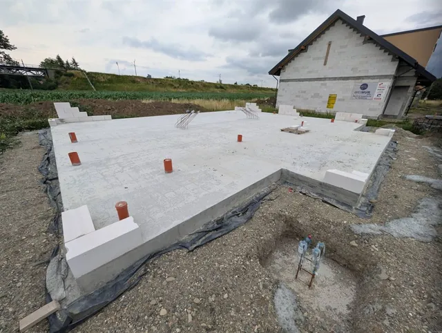 Płyta fundamentowa z zarysowanym podcieniem wejściowym i pionami kanalizacji, w tle gotowy stan surowy sąsiedniego budynku