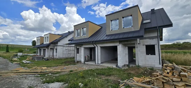 Dwa budynki dwulokalowe od strony podjazdów — gotowe dachy z blachy na rąbek stojący, lukarny z oknami i rynny, przy drugim rusztowanie